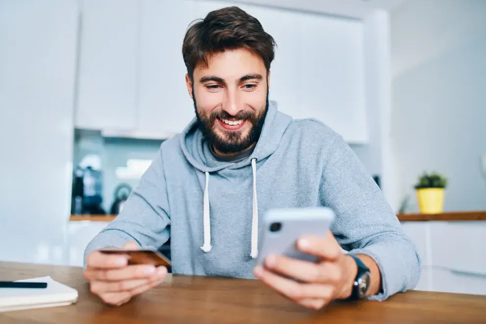 smiling young man sitting at home counter and paying online with credit card and smartphone smiling young man sitting at home counter and paying online with credit card and smartphone